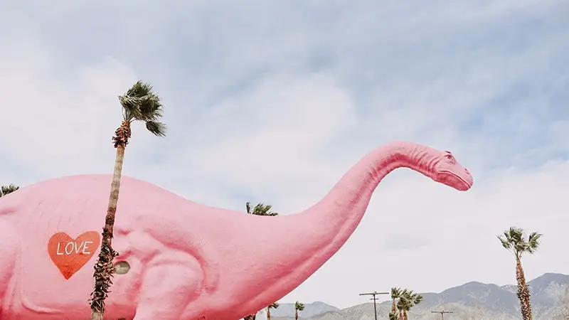 A massive pink dinosaur statue stands under a partly cloudy sky, flanked by several palm trees. A red heart with the word "LOVE" painted inside adorns its side. Mountains loom in the distance—a perfect spot to explore Palm Springs and one of the best places to visit for unique activities.