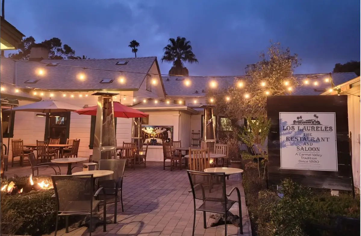 An outdoor restaurant patio at dusk, decorated with string lights glowing warmly above. The space features several tables and chairs, an overhead heater, and a fire pit. A sign reads "Los Laureles Restaurant and Saloon," part of Carmel Group Hotels. The sky is cloudy and the area is peaceful.