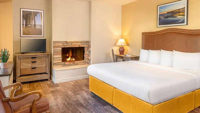 A cozy hotel room with a large bed featuring white linens and a mustard-yellow base. The room, reminiscent of Southern California Missions, has a lit fireplace, a TV on a wooden dresser, an armchair, and a small desk with a lamp. Art adorns the walls, and the space is warmly lit and inviting.