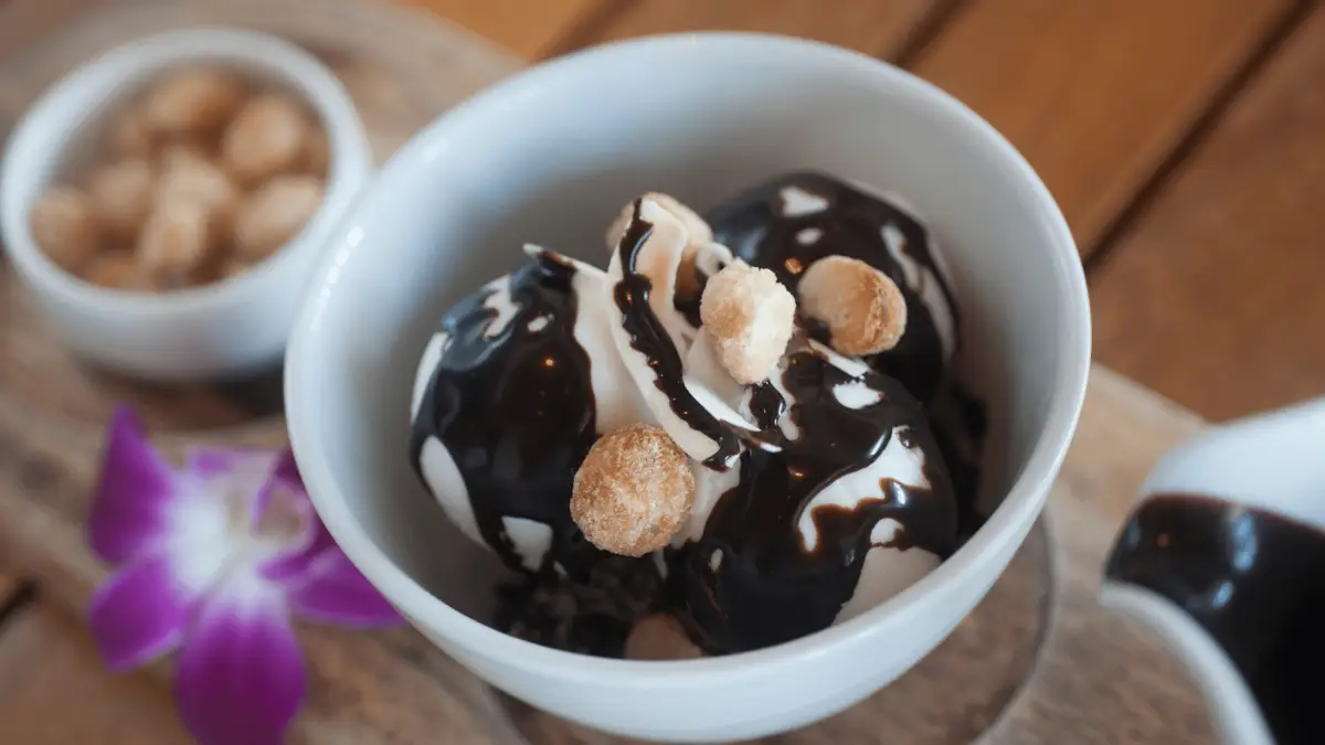 A white bowl with vanilla ice cream drizzled with chocolate sauce and topped with small biscuit-like treats. A purple orchid is placed beside the bowl. In the background, another small bowl filled with peanuts is visible on a wooden surface, showcasing one of the best desserts Maui Wailea has to offer.