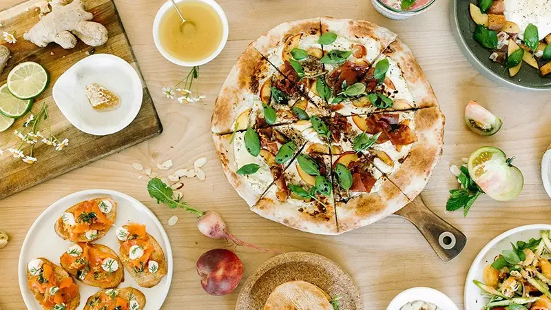 A wooden table is set with a variety of dishes including a freshly baked pizza topped with greens, salmon-topped bruschetta, a bowl of honey, sliced fruits, and a bowl of salad. The table, reminiscent of Southern California Missions' vibrant gatherings, is garnished with flowers and lime slices.