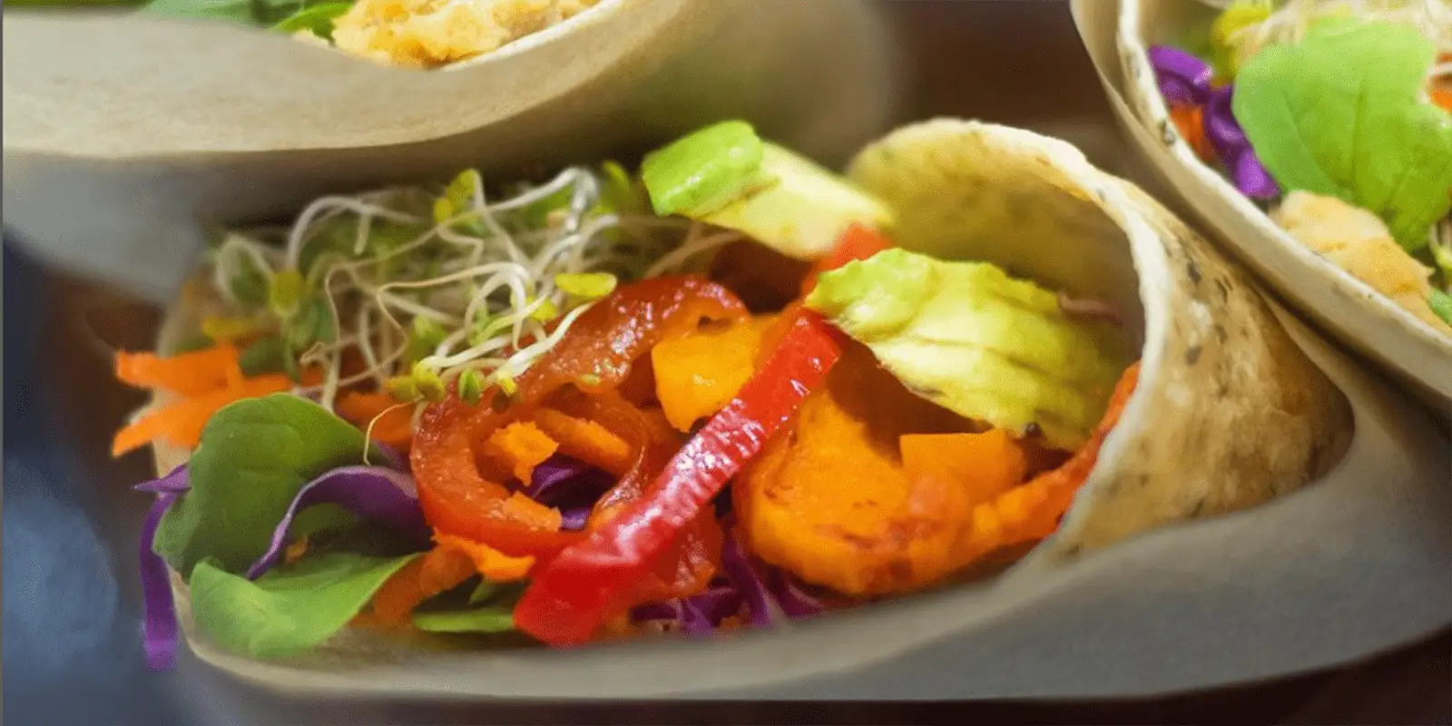 A close-up of a colorful, fresh veggie taco, the best lunch on the North Shore. The taco is filled with vibrant ingredients including sliced avocado, red bell peppers, shredded purple cabbage, carrots, greens, and alfalfa sprouts, and wrapped in a wheat tortilla.