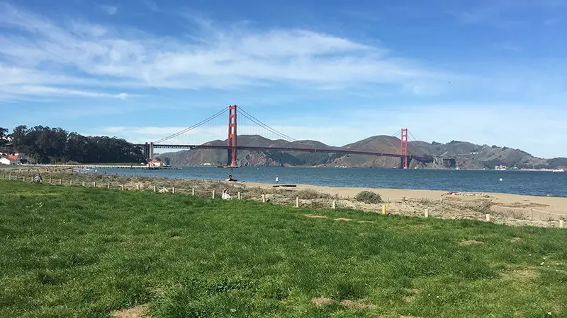 A scenic view of the Golden Gate Bridge in San Francisco, spanning across the water with hills in the background. The foreground features a grassy area and a sandy beach, with a person in the distance enjoying the landscape on a clear, sunny day—one of Five Ways to Celebrate July 4th in the Bay Area.