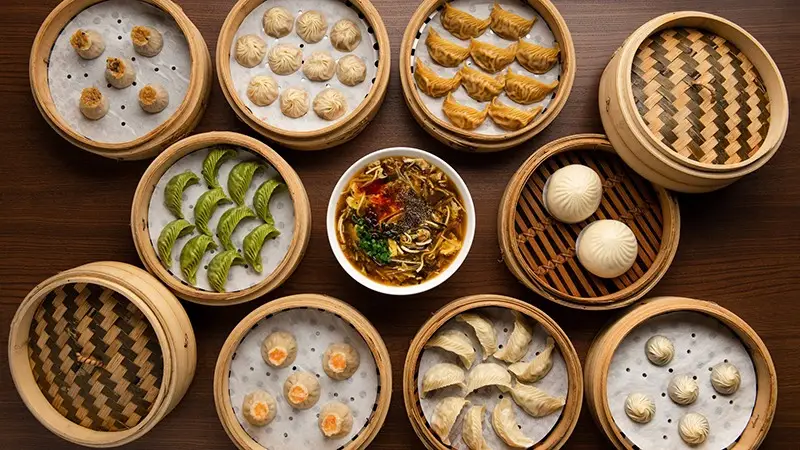 An overhead view of various bamboo steamers filled with different types of dumplings and buns, arranged in a circular formation around a bowl of soup. The vibrant assortment of dim sum resembles the diverse and historic charm found in Southern California Missions.
