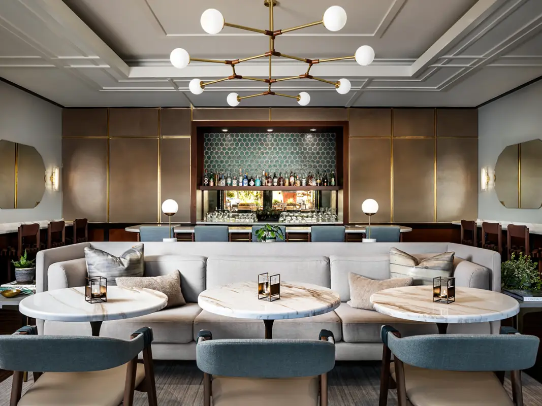 A contemporary lounge room with a central beige couch, round marble-topped tables, and cushioned chairs. In the background, there's a well-stocked bar with a hexagonal tile backsplash and modern light fixtures. A large, modern chandelier hangs from the ceiling in this California Eco Hotel's eco-friendly design.