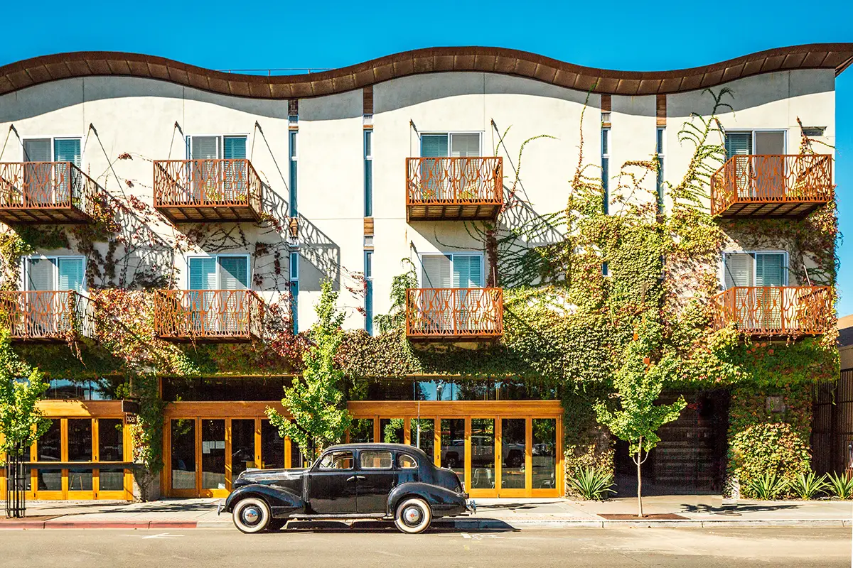 A vintage black car is parked in front of a modern, three-story building with a wavy roofline, reminiscent of California Eco hotels. The building features multiple balconies with wooden railings, and parts of the facade are covered with climbing vines. The clear blue sky completes this eco-friendly scene.