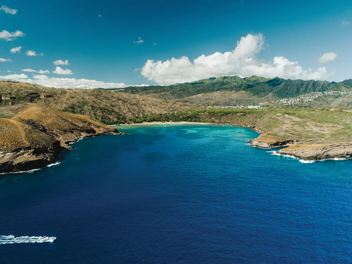 Aerial view of a scenic bay in Oahu with turquoise waters surrounded by rocky cliffs and green hills under a bright blue sky with scattered white clouds. Dense green foliage covers the hills in the background, and small white structures are visible in the distance, hinting at vibrant marine life below.