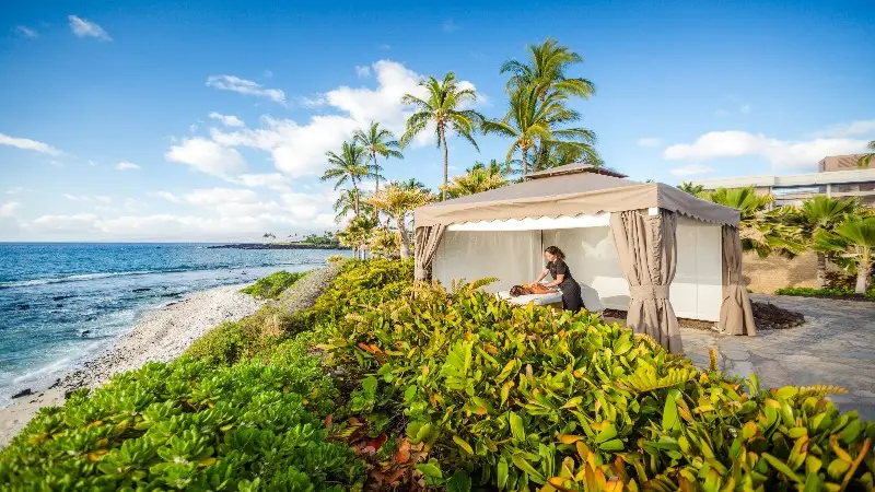 A person receives a massage in an open-air cabana beside a scenic shoreline. The setting includes lush greenery, palm trees, and a rocky beach with calm, blue ocean waters under a partly cloudy sky.
