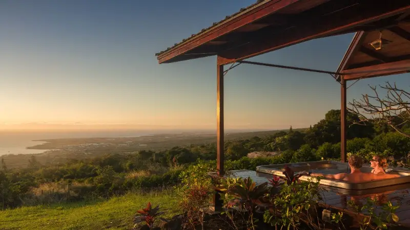 Two people relax in an outdoor hot tub under a wooden gazebo, overlooking a scenic view of lush greenery, a coastal town, and the ocean with a clear sky at sunset. The atmosphere is tranquil, and the setting sun casts a warm glow over the landscape—truly one of the best romantic hotels on Big Island.