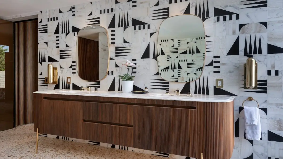 Modern bathroom featuring a large wooden vanity with a sleek white countertop and twin oval mirrors. The wall is decorated with black and white geometric tiles. Brass accents, including faucets, light fixtures, and a towel holder, add a touch of sophistication reminiscent of the best hotels in Palm Springs.