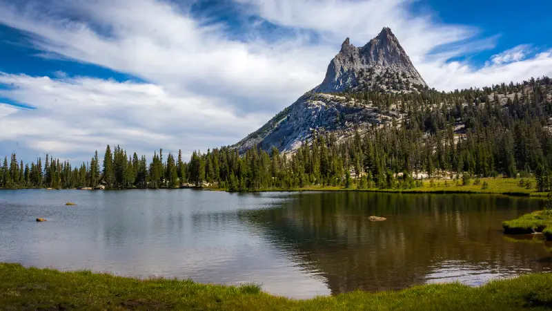 A serene lake reflects a majestic, jagged mountain peak under a partly cloudy blue sky. The landscape features dense, green forests around the water's edge and a grassy shoreline in the foreground. This scenic view along the John Muir Trail epitomizes tranquility in the wilderness.