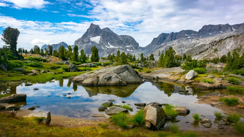 A tranquil mountain scene with clear skies, featuring a serene lake reflecting towering rocky peaks in the background, evokes the spirit of the John Muir Trail. Greenery surrounds the lake, with scattered boulders and patches of grass contributing to the picturesque landscape.