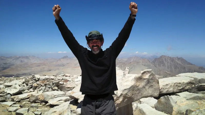 A person stands atop a rocky mountain peak on the John Muir Trail, arms raised in triumph. They are smiling widely, wearing a black long-sleeved shirt, a green hat, and dark pants. The background shows a vast mountainous landscape under a clear, blue sky.