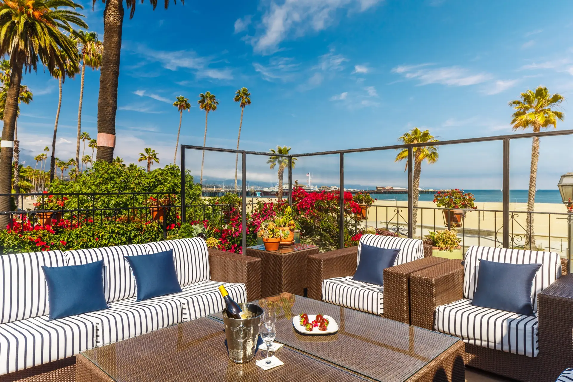 A luxurious outdoor patio in one of the best group getaway hotels Santa Barbara has to offer, featuring wicker furniture adorned with striped cushions and blue accent pillows. A glass-top coffee table displays a metal ice bucket, glasses, and a plate of snacks. Vibrant flowers, palm trees, and the ocean are visible in the background.
