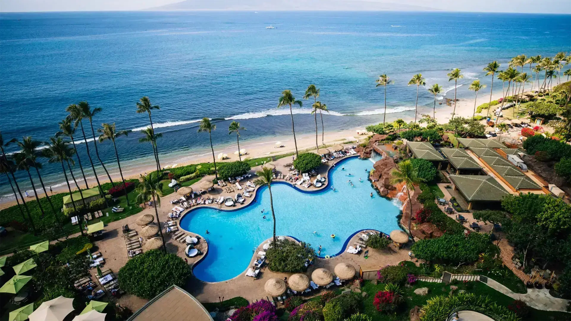 Aerial view of one of the best family hotels in Maui, featuring a large, uniquely shaped swimming pool surrounded by lounge chairs and lush greenery. The resort is bordered by a sandy beach and clear turquoise ocean waves, with palm trees scattered throughout the landscape.