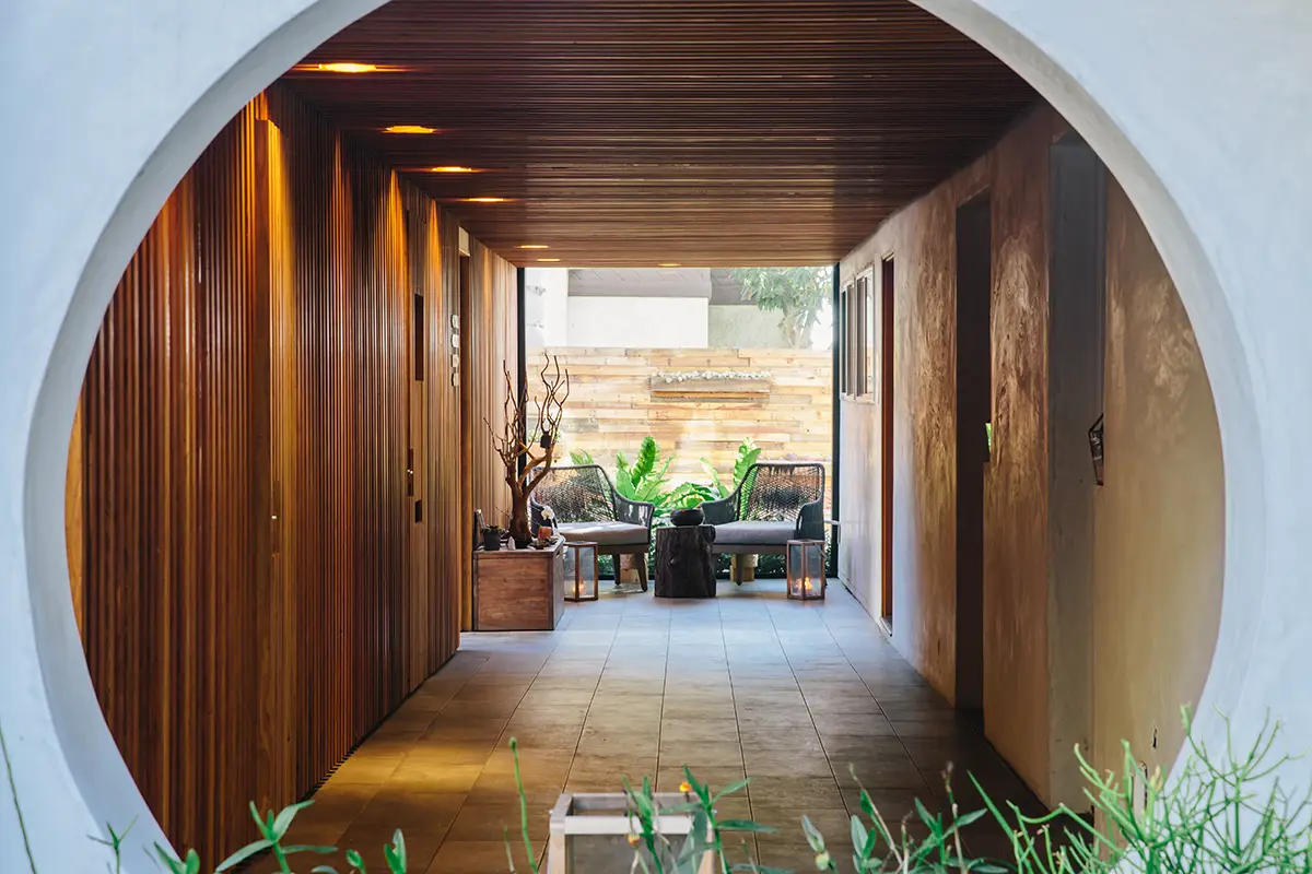 A modern hallway with wooden panel walls and slate flooring leads to an outdoor seating area. The round entrance frames a cozy patio with wicker chairs, potted plants, and a wooden fence in the background, creating an eco-friendly blend of indoor and outdoor spaces reminiscent of California eco hotels.