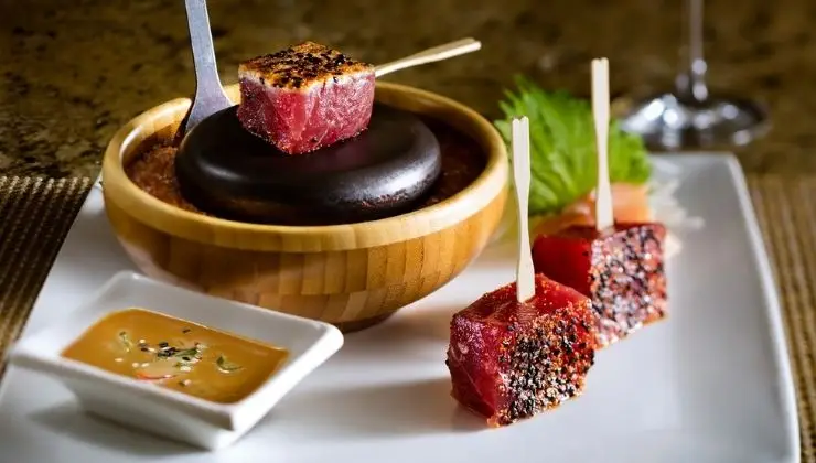 A dish featuring skewered, seared tuna cubes with sesame seeds, served on a small wooden bowl containing a heated stone. Alongside, there is a dipping sauce in a small square dish and garnishes on a white plate. Experience one of the best dinners Kihei Wailea has to offer!