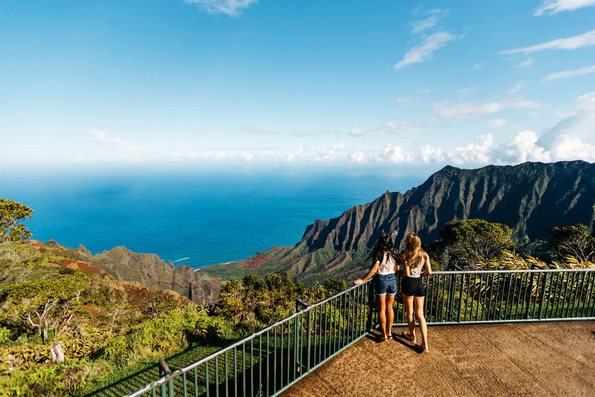 Two people stand at a railing overlooking a scenic view of lush green mountains and the expansive blue ocean. The sky is clear with scattered clouds, and the sunlight illuminates the landscape, highlighting the vibrant natural colors, while Kauai's sealife thrives just below the surface.