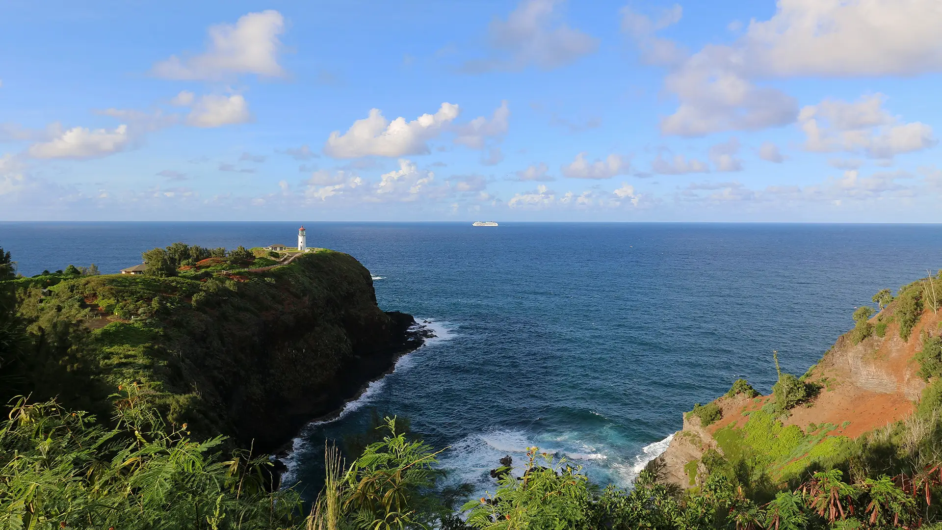 A scenic view of the ocean with rocky cliffs in the foreground, lush green vegetation, and a distant lighthouse perched on the edge of the cliff. The sky is blue with scattered clouds, and a ship can be seen on the horizon—certainly one of the best places to visit on Kauai.