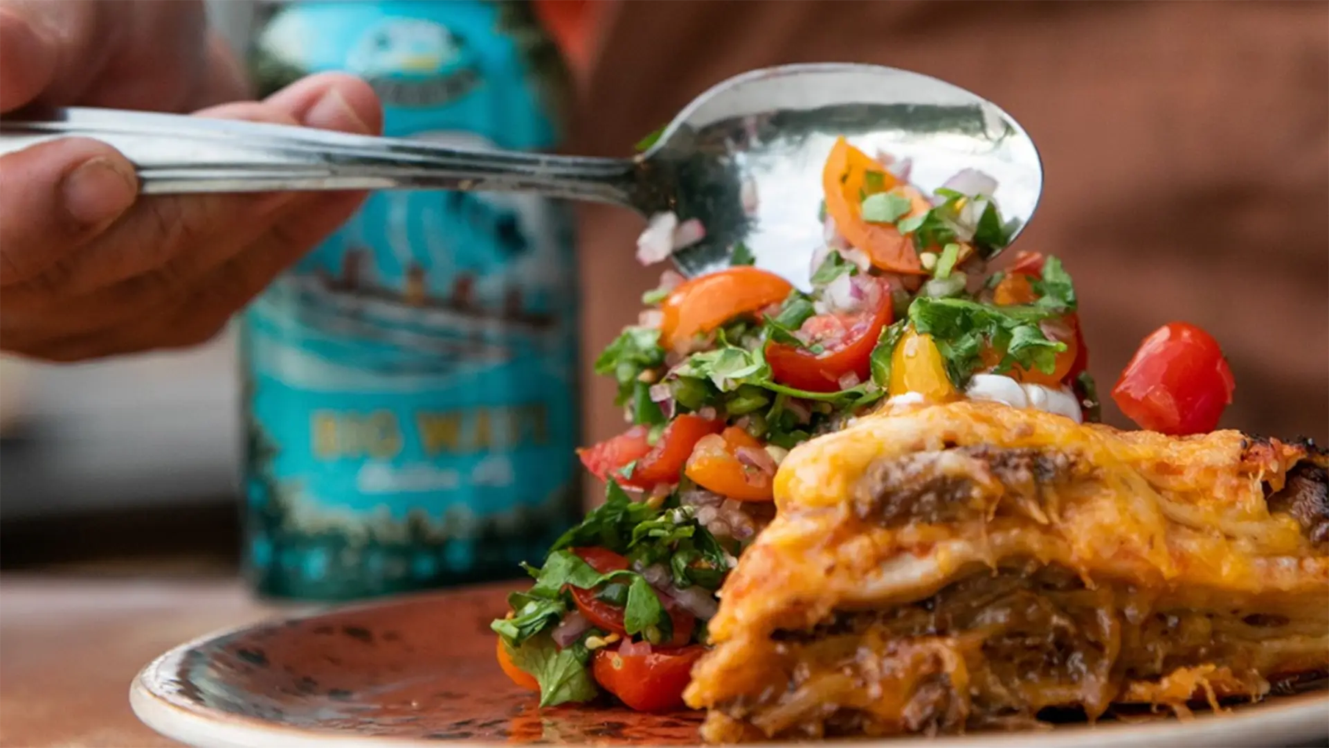 Close-up of a person holding a spoon over a plate of food. The plate contains the best dinner in Kona—a layered dish with melted cheese and meat, accompanied by a colorful vegetable salad with tomatoes, onions, and herbs. A can of Big Wave Golden Ale beer is visible in the background.