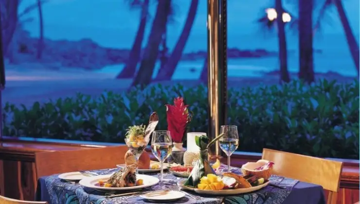 A dining table is set with various dishes, including seafood and fruit, surrounded by wine glasses. The backdrop features an evening beach scene from Paia and Hana Maui, with palm trees and a deep blue sky, viewed through a large window. The table has an elegant tablecloth.
