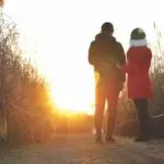 Two people, dressed warmly, stand side by side on a path flanked by tall grasses in Marin, looking towards the sunset. One wears a black jacket and the other a red coat with white fur trim. The golden sunlight bathes the scene, creating a warm, serene atmosphere perfect for discussing health tips.