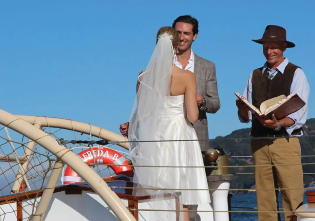 A couple is getting married on a boat, one of Marin's Top Wedding locations. The bride, in a white dress and veil, faces away from the camera, holding hands with the groom in a gray suit. The officiant, wearing a hat and vest, stands beside them holding an open book. It's a sunny day with clear blue sky.
