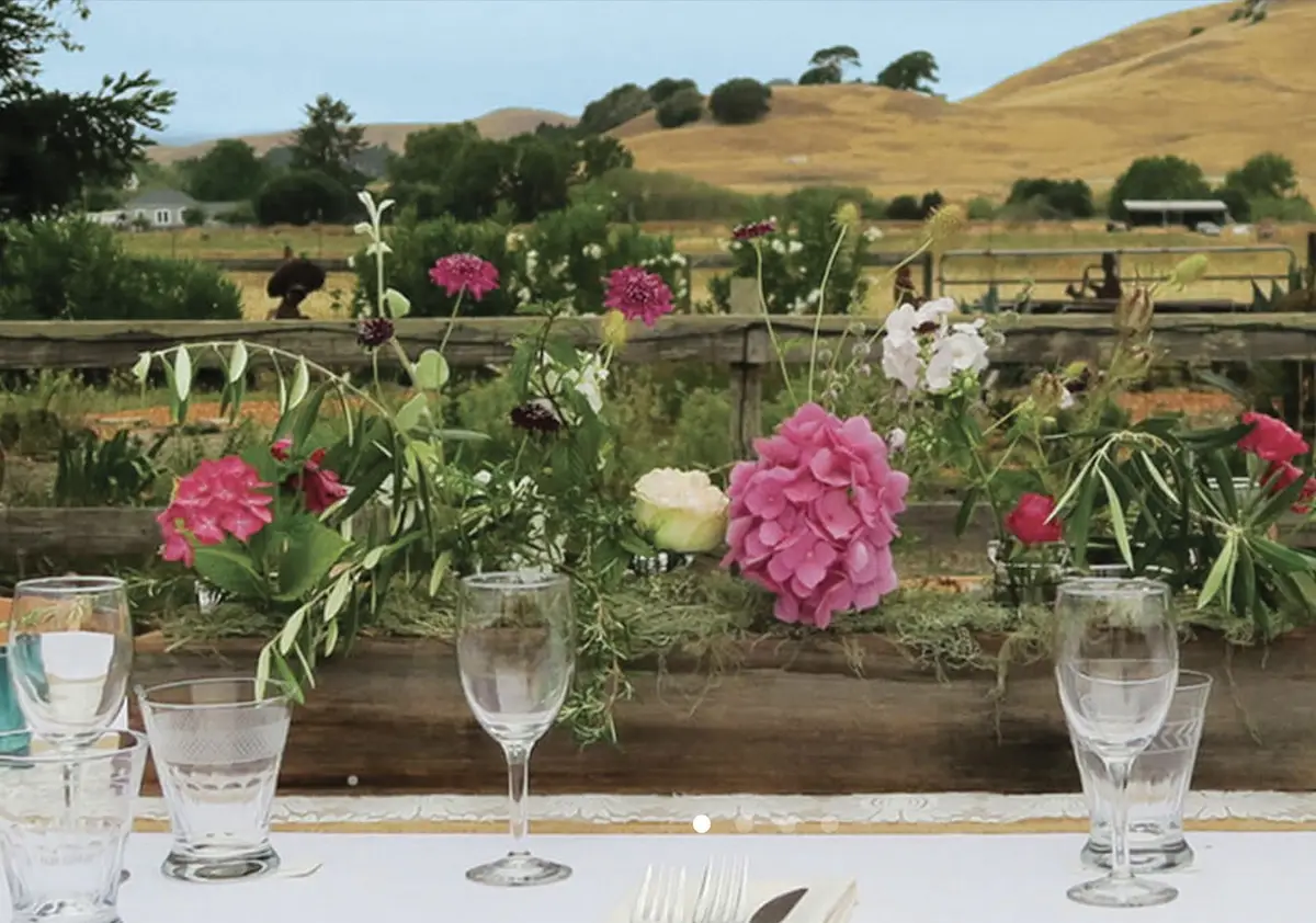 A rustic outdoor dining setup, one of Marin's top wedding locations, features a floral arrangement with pink, purple, and white flowers decorating a wooden fence in the background. White tablecloth-covered tables are set with glasses, plates, and cutlery. Rolling hills and trees are visible in the distance.