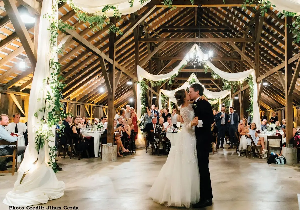 A newlywed couple shares their first dance in a rustic barn, one of Marin's top wedding locations, decorated with draped white fabric and greenery. Guests seated at round tables surround the dance floor, watching the couple. The wooden ceiling is adorned with string lights, creating a warm, festive atmosphere.