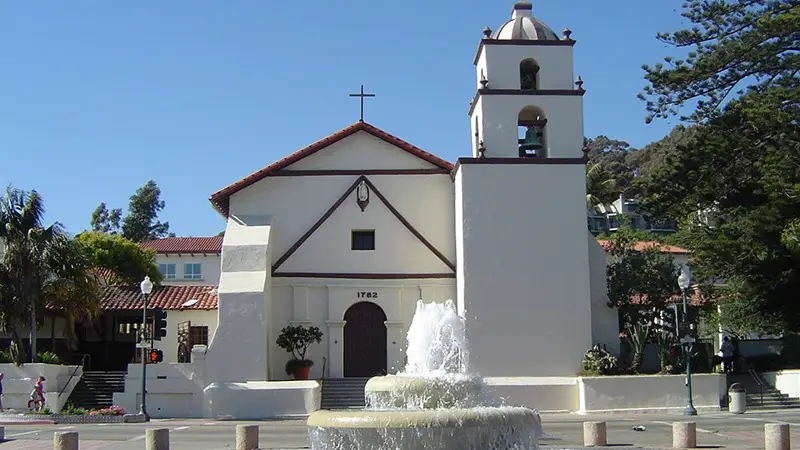 A historic white church with a red-tiled roof and a bell tower stands prominently under a clear blue sky, reminiscent of the Southern California Missions. A cross adorns the top of the building, and a fountain in the foreground enhances the picturesque setting. Trees and greenery surround the area.