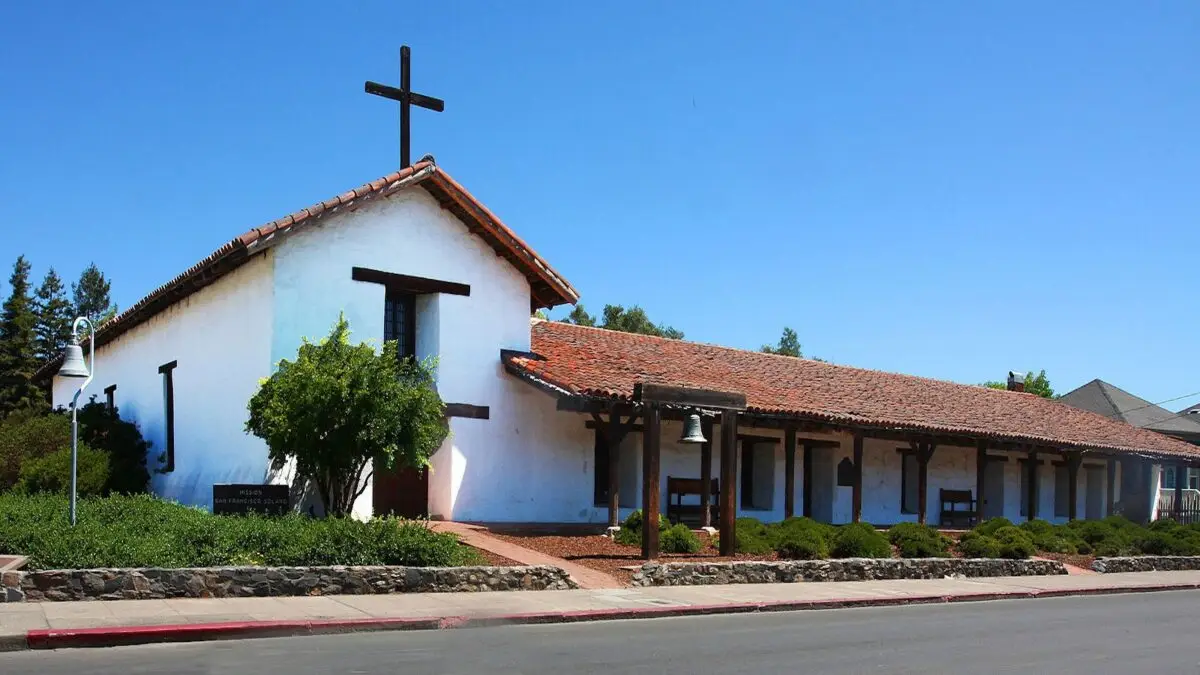 A historic mission-style building with white stucco walls and a tile roof stands under a clear blue sky in one of the best places to visit in Sonoma County. A large wooden cross is fixed atop the roof, and there is a small bell in the courtyard, surrounded by well-maintained shrubs.