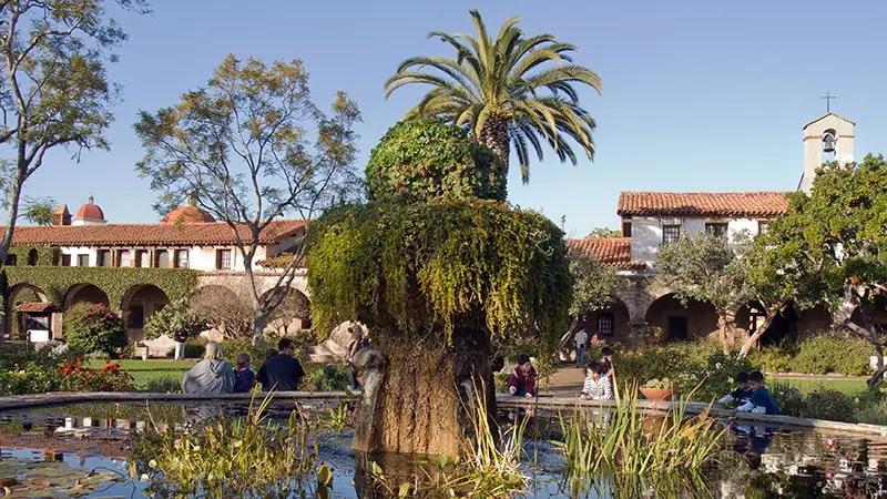 Scenic view of a historic mission courtyard with a lush garden and a large stone fountain covered in greenery at the center. The background features Mediterranean-style buildings with red-tile roofs and archways, typical of Southern California Missions, along with palm and deciduous trees under a clear blue sky.