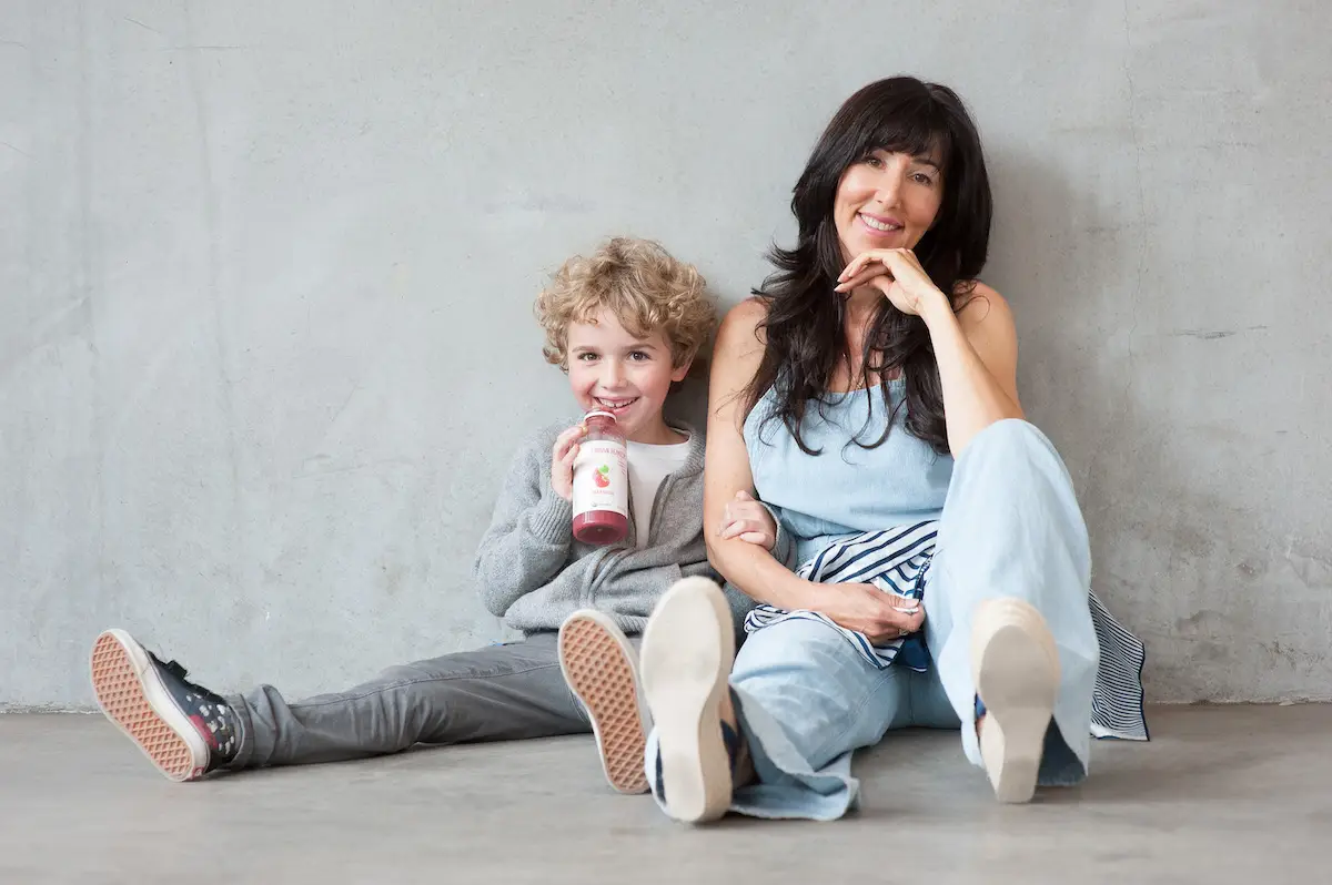 A woman and a child sit on the floor against a plain wall. The woman, with dark hair and wearing light blue clothing, smiles and rests her chin on her hand. The child, with curly blond hair, drinks from an Urban Remedy bottle and leans against the woman, also smiling.
