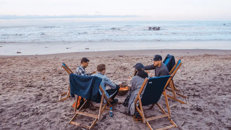 A group of four people sits on beach chairs around a fire pit on the sand at Half Moon Bay, facing the ocean. They appear to be engaged in conversation. The sky is clear, and the waves are gently crashing onto the shore in the background— a perfect coastal getaway.