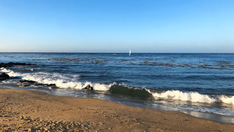 A serene beach scene with gentle waves lapping against the sandy shore, among The 7 Natural Wonders of California. The sky is clear with a bright blue hue, and a lone white sailboat is seen on the horizon, sailing across the calm sea. Rocks are scattered near the shoreline.
