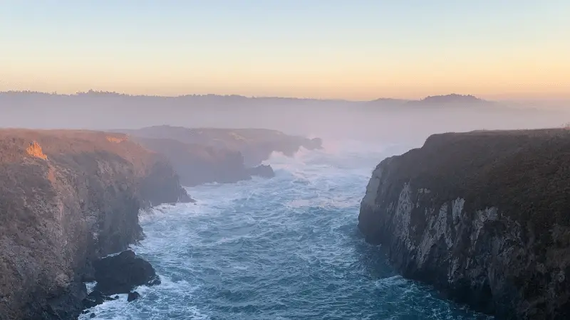 A picturesque coastal scene at dusk, featuring rugged cliffs and choppy ocean waves crashing against the rocks. The horizon is shrouded in mist, with the evening sun casting a soft, golden hue over the landscape of Northern California's small town getaways, creating a serene and ethereal atmosphere.