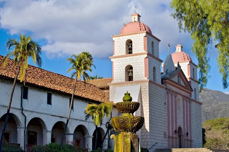 A historic mission stands under a partly cloudy sky, featuring white stone walls, red-tiled roofs, and two bell towers with pink domes. Palm trees and a leafy tree frame the scene while a tiered stone fountain is prominent in the foreground—one of the best things to do in Santa Barbara.