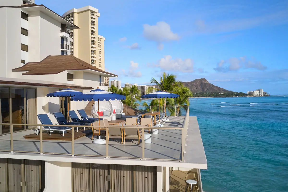 Rooftop lounge area with sun loungers and tables under blue umbrellas, overlooking a pristine ocean teeming with Oahu marine life and distant mountains. Tall buildings nearby, clear blue sky, and palm trees add to the tropical ambiance.