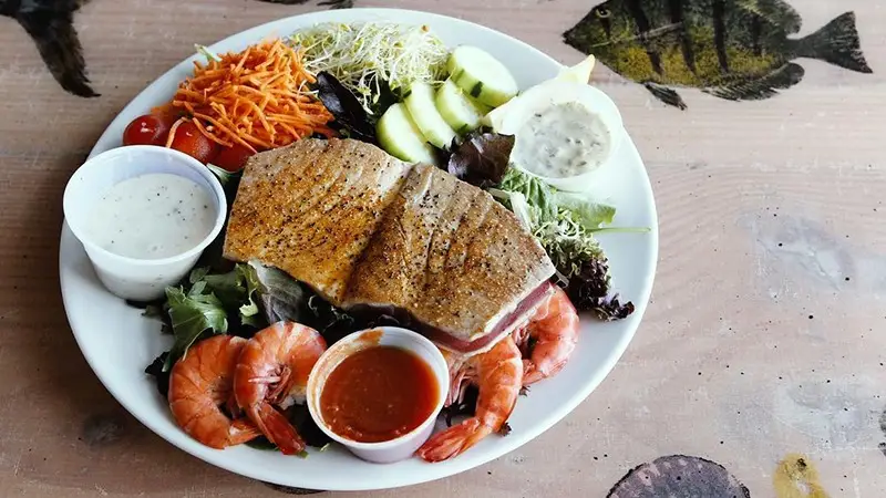 A colorful dinner salad featuring pieces of grilled fish, shrimp, mixed greens, shredded carrots, cherry tomatoes, cucumber slices, and two dressing cups is served on a white plate. The table has a rustic look with fish illustrations that echo the charm of Paia and Hana in Maui.