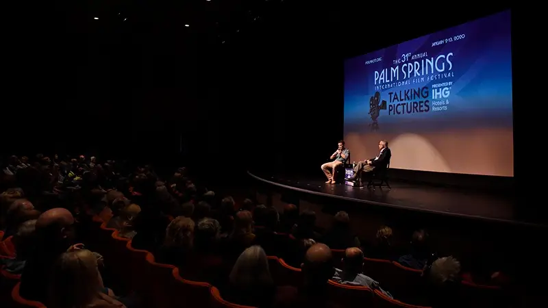 A seated audience in a dimly lit auditorium listens to two people on stage during a conversation event at the 31st Palm Springs International Film Festival. The stage backdrop, echoing "The Ultimate Guide to Palm Springs," displays the festival's title and event details under a "Talking Pictures" banner.