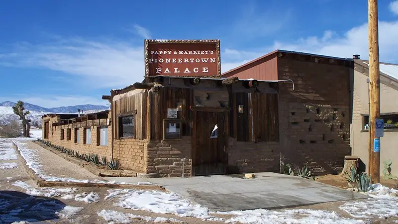 A rustic, Western-style building with a sign that reads "Pappy & Harriet's Pioneertown Palace." The structure is made of wood and brick, set against a desert landscape with some snow patches. With a clear blue sky overhead, it's one of the best places to visit near Palm Springs for those looking to explore.