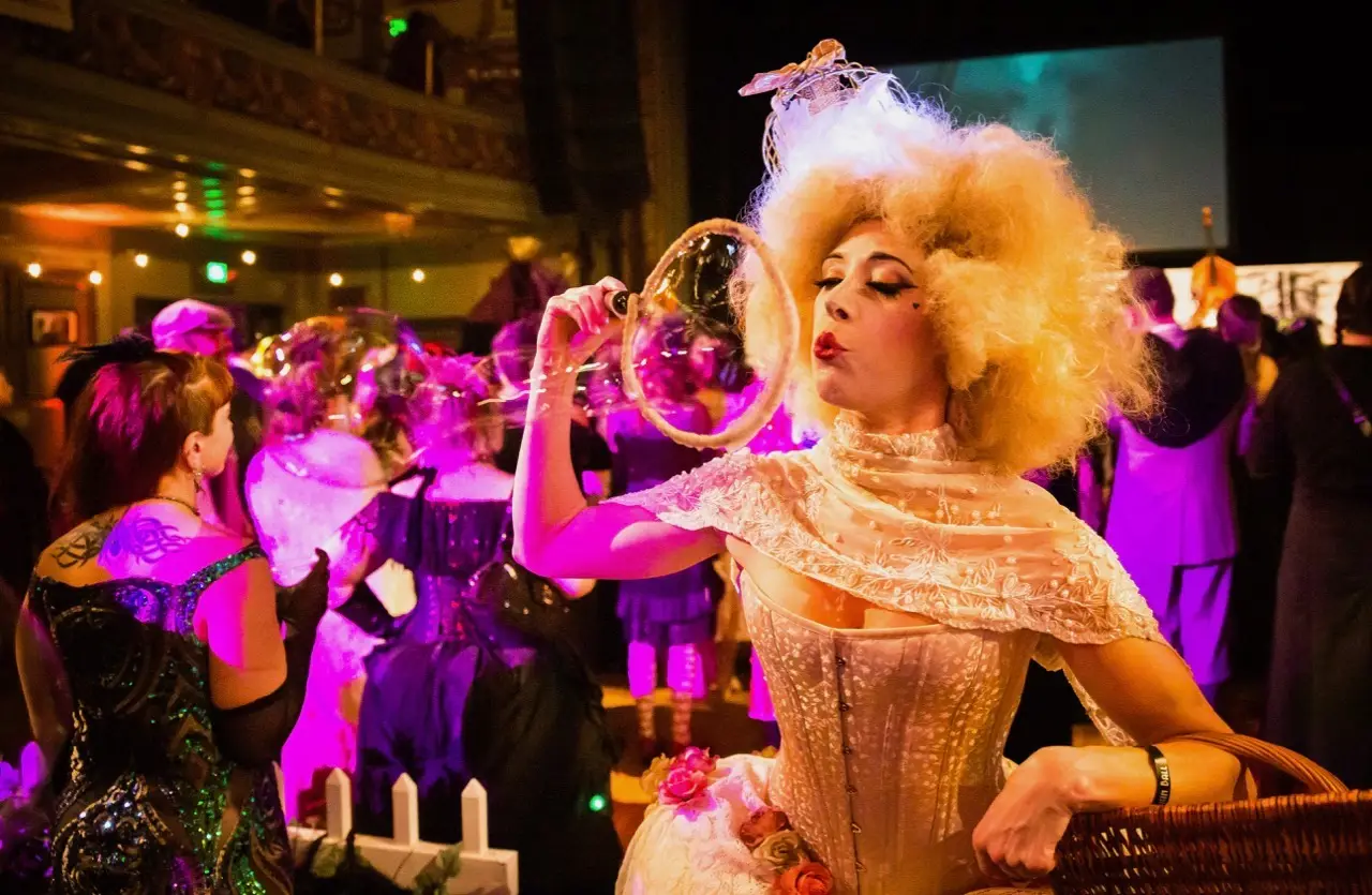 A woman in a flamboyant, vintage costume blows bubbles at an event with vibrant lighting. She has blonde, curly hair and wears an ornate dress. People in elegant attire surround her, creating a festive atmosphere in a large, decorated hall—one of the top things to do in the California Bay Area this January.