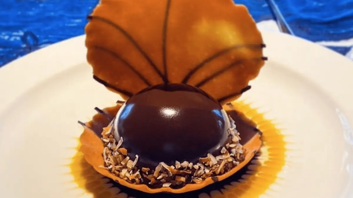 A sophisticated dessert featuring a glossy, dark chocolate dome garnished with toasted coconut flakes and seated in a caramelized shell on a white plate. The backdrop hints at a blue, textured surface, adding a luxurious touch to the presentation. Truly among the best desserts Maui Wailea offers.