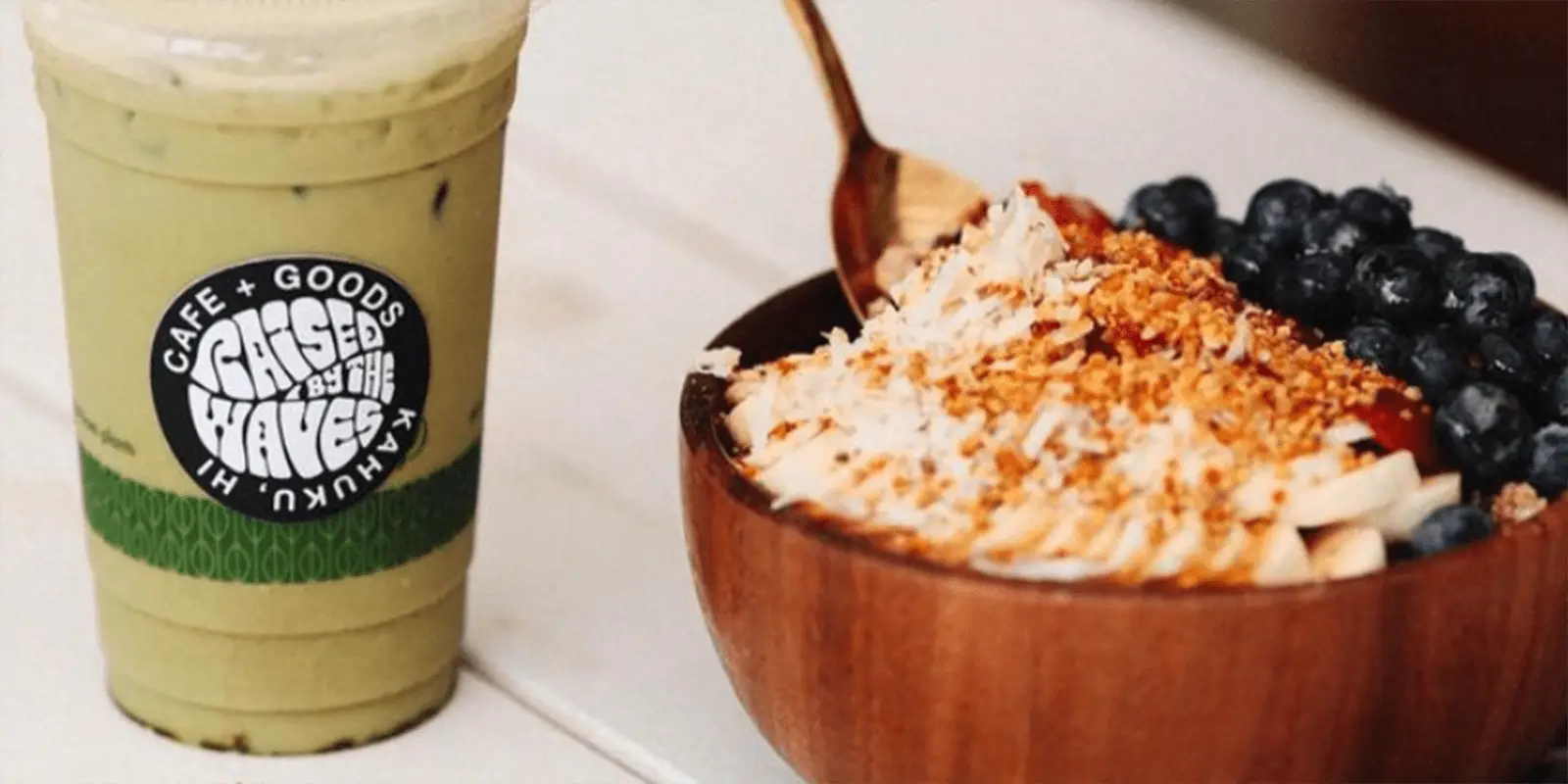 A wooden bowl filled with an açai bowl topped with banana slices, blueberries, shredded coconut, granola, and seeds. A fork rests on the edge of the best lunch North Shore has to offer. Next to it is a green iced matcha latte in a plastic cup with the logo "Café + Goods" on the sleeve.