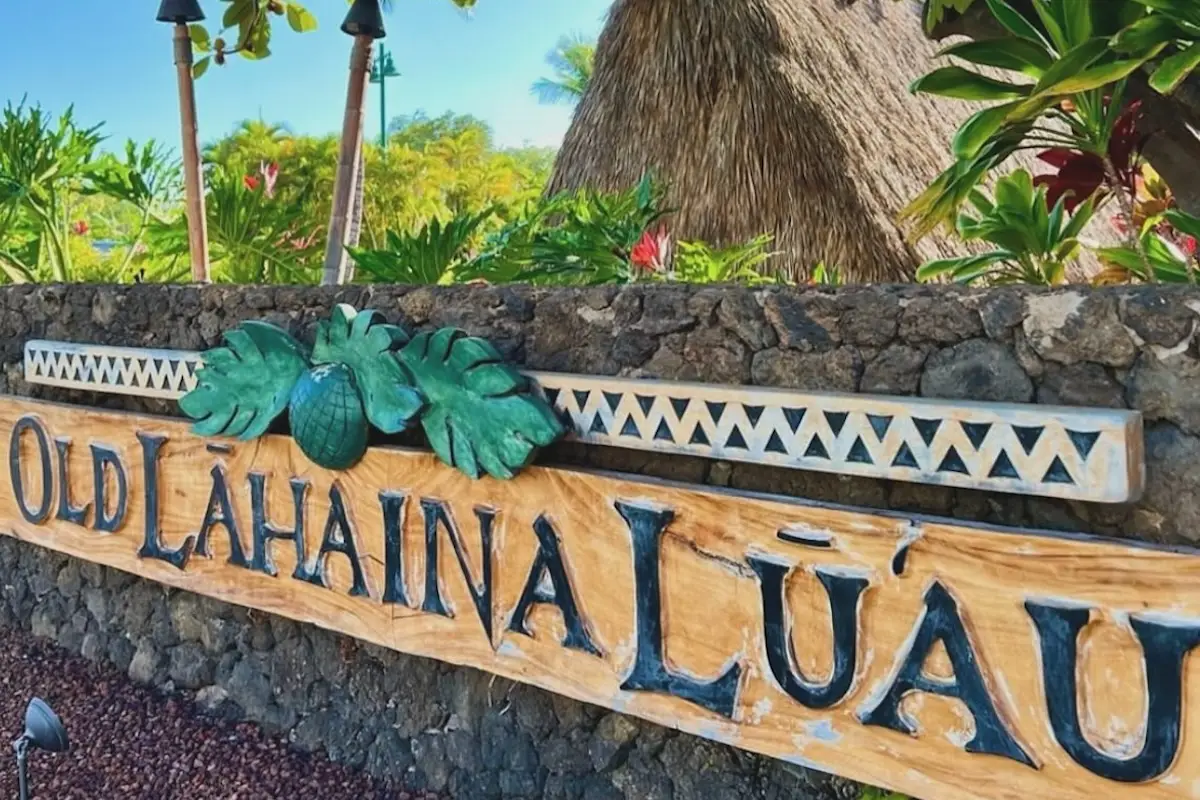 A wooden sign with the words "OLD LAHAINA LUAU" inscribed in large, black letters is mounted on a stone wall. The sign is adorned with green tropical leaves and a pineapple, capturing the essence of West Maui. Lush vegetation and tiki huts are visible in the background.