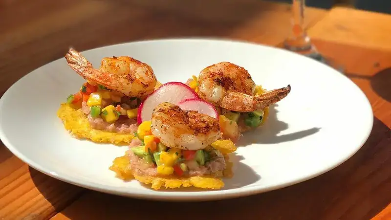 A white plate with three shrimp appetizers is on a wooden table. Each appetizer consists of a grilled shrimp placed on a bed of colorful vegetable salsa atop a crispy base, garnished with radish slices. Sunlight, reminiscent of the warmth found near Southern California Missions, casts soft shadows on the dish and table.