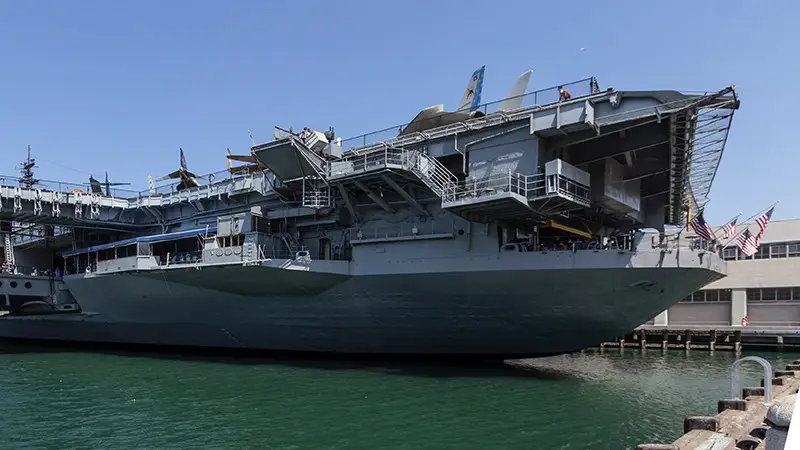 A large gray aircraft carrier docked at a port, reminiscent of the grandeur seen in Southern California missions. Several aircraft are on the deck, with an American flag towards the rear. The green water, part of the pier, and a dockside building are visible under a clear sky.