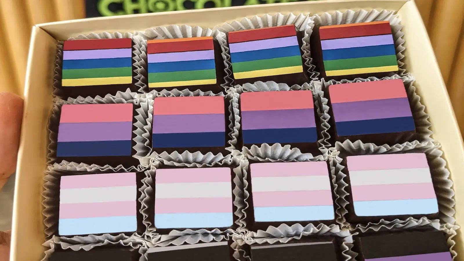 Celebrate San Francisco Pride all month long with a box of chocolates, each adorned with different LGBTQ+ pride flags, including rainbow, bisexual, lesbian, non-binary, and other pride stripes. The chocolates are neatly arranged in rows with paper wrappers separating them.