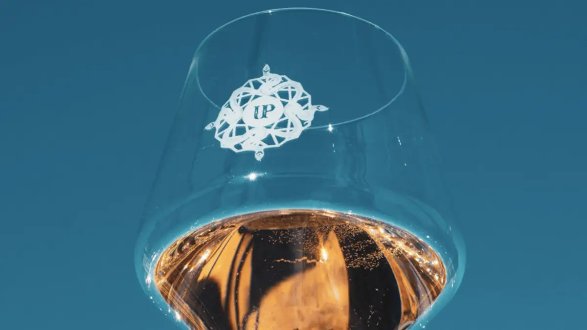 Close-up photograph of a clear wine glass filled with rosé wine against a clear blue sky. The glass features an elegant, white logo with the letters "I" and "P" at the center. The light pink wine, reminiscent of Provence, has bubbles near the surface. Perfect for International Rosé Day.