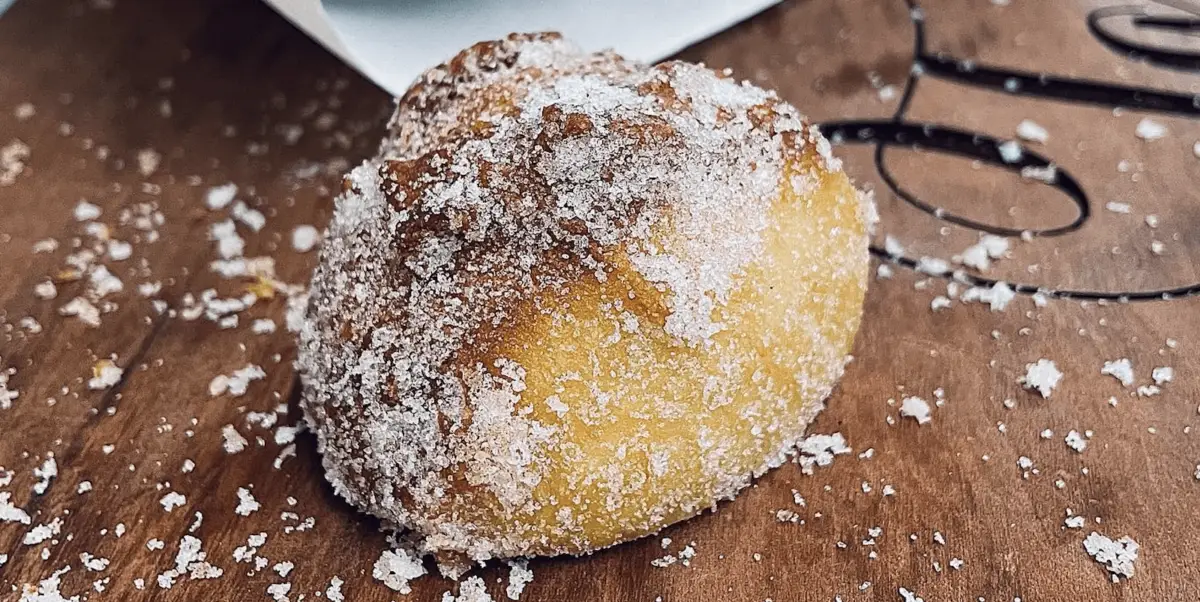 A sugar-coated, golden-brown pastry sits on a wooden surface. The pastry appears soft and fluffy, with a light dusting of sugar granules on top and around it. Truly one of the best desserts you'll find in Maui Wailea.